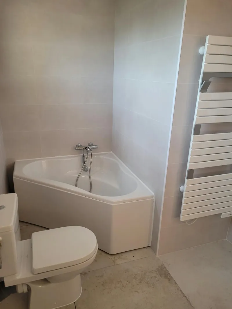 La même salle de bain vue de face : baignoire d’angle, WC et sèche-serviettes